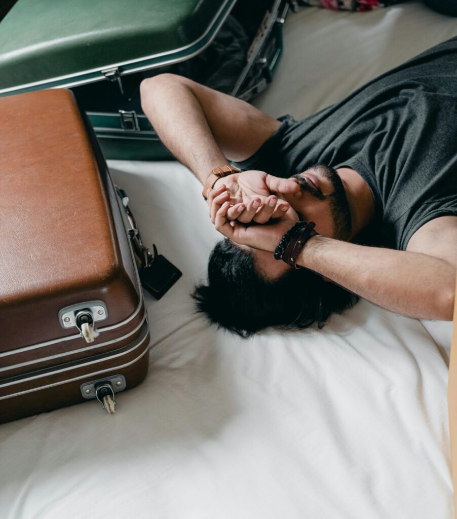 A person lies on a bed, covering their face with their hands, looking exhausted. Next to them, two vintage suitcases—one green and one brown—are slightly open, suggesting travel or packing stress.